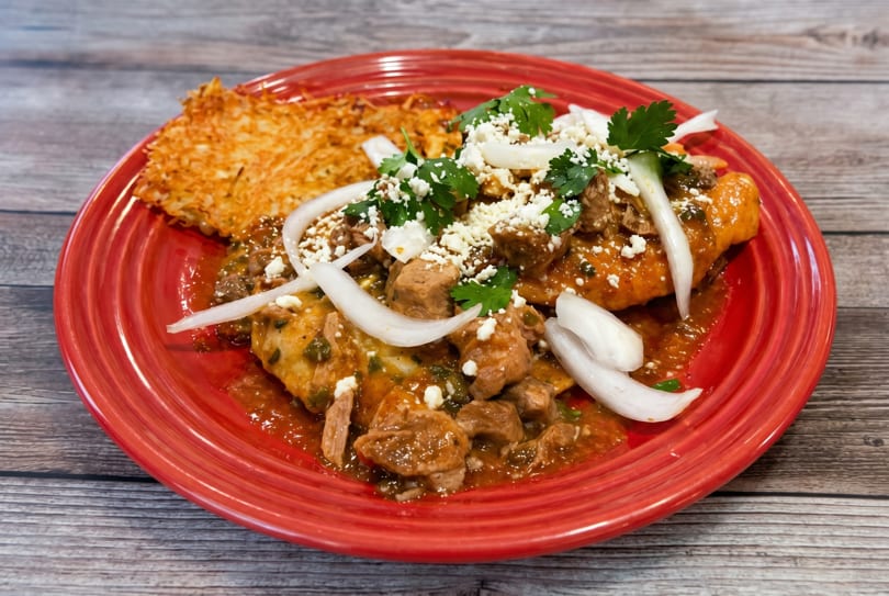 Savory pork stew with onions, cilantro, and cheese, served with a crispy hash brown on a red plate.