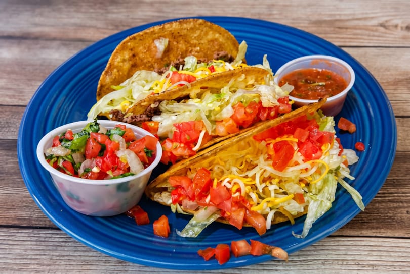 Three hard-shell tacos filled with meat, lettuce, cheese, and tomatoes, served with salsa and pico de gallo.