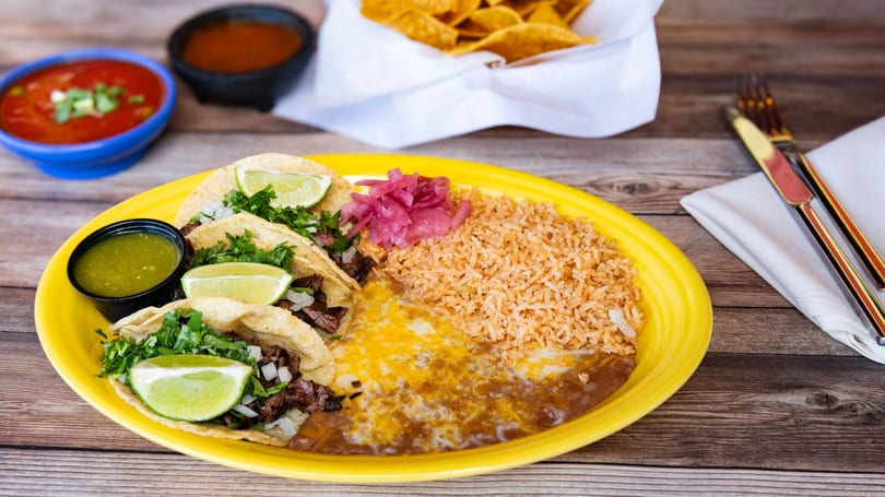Three street tacos with lime, cilantro, rice, beans, and pickled onions on a yellow plate.