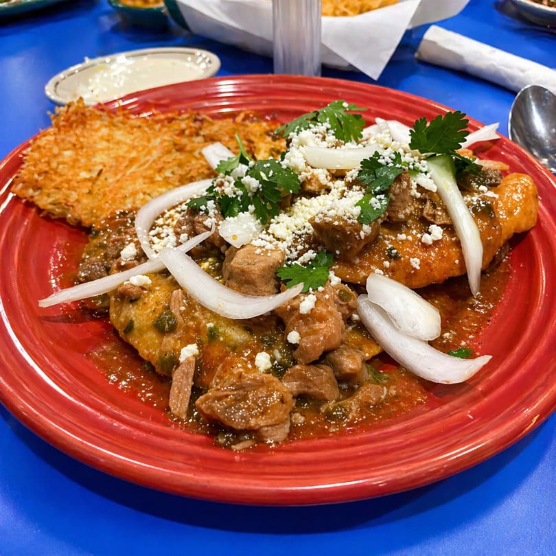 Red plate of chilaquiles with pork, cheese, onions, cilantro, and a hash brown.