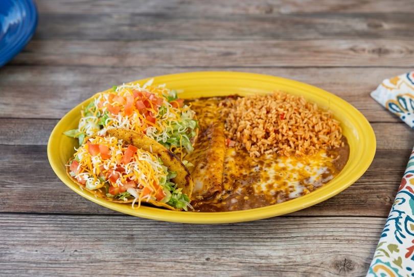 A yellow plate holds two tacos, enchiladas, refried beans, and rice on a wooden table.