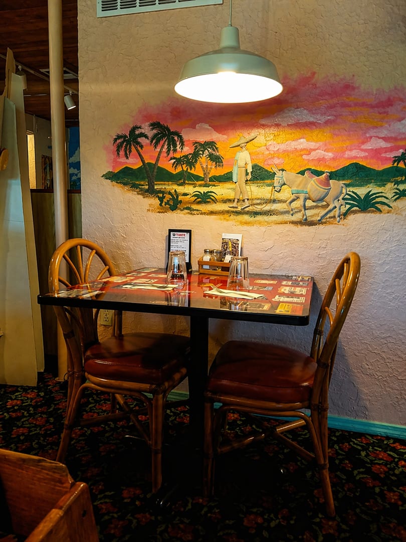 A restaurant table set for two, with a colorful mural of a desert sunset and palm trees in the background.