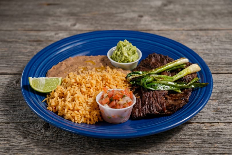 Carne asada with grilled green onions, rice, refried beans, guacamole, pico de gallo, and a lime wedge.